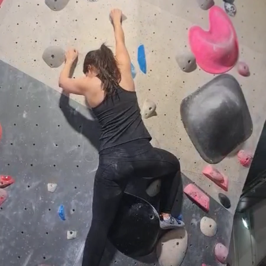 Natalie bouldering indoors, reaching for a hold high on an overhanging wall