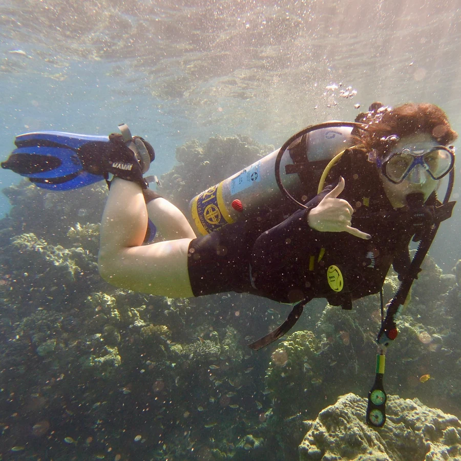 Natalie scuba diving over a coral reef, throwing a shaka sign at the camera
