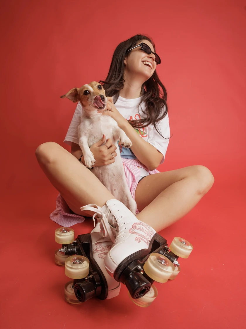 Natalie sitting cross-legged on a red backdrop in roller skates and sunglasses, laughing, holding a small dog